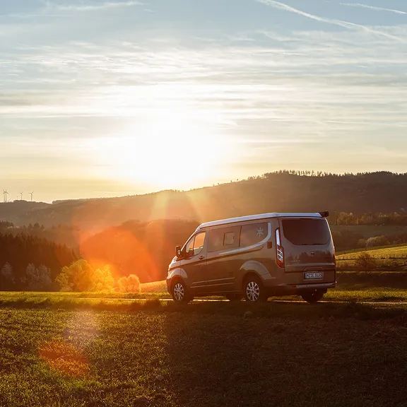 Karmann-Mobil Campingbus fährt auf einer Straße mit weitem Blick über bewaldete Hügel bei stimmungsvollem Sonnenuntergang.